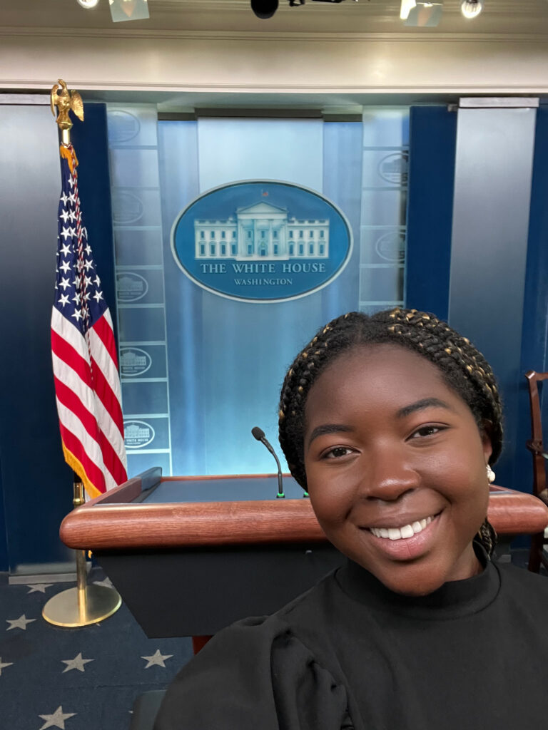 Daniella smiling in the White House Press Room, with a podium and American flag in the background.