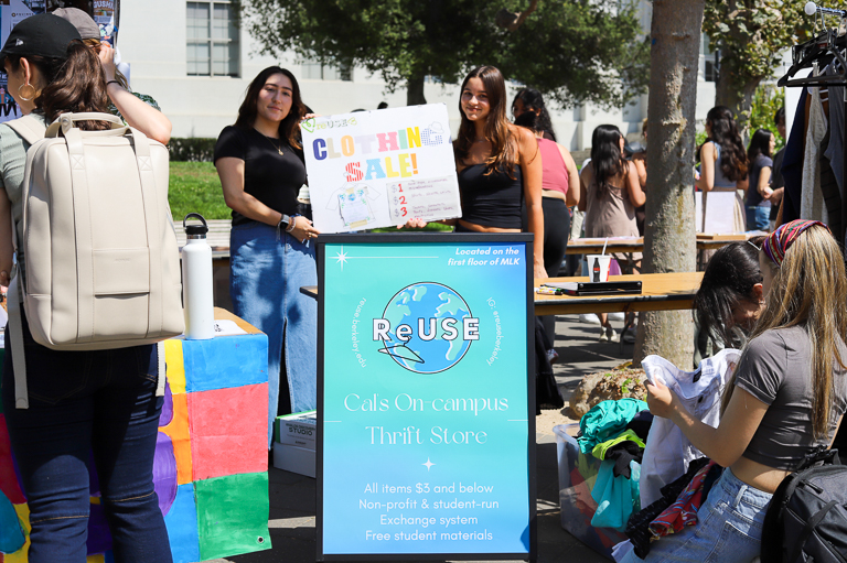 Two members of the ReUSE club hold up a sign reading "clothing sale!" In front of their table is a blue sandwich board that describes ReUSE as a on-campus thrift store and clothing sign. 