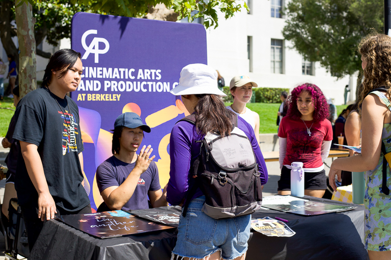 Jarvis Nguyen, Steven Zeng, Addie Tweet, and one other member of the Cinematic Arts & Production at UC Berkeley speak to a prospective club member while tabling. There is a colorful sign displaying Cinematic Arts & Production Club at Berkeley behind the club members.