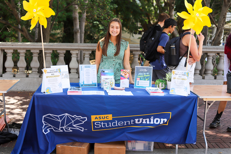 Jill Matson smiles wearing a green dress while tabling for the ASUC Student Union at Calapalooza. The ASUC Student Union banner is dark blue, and yellow pinwheels rest atop it.