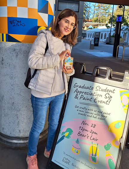 Marjan holds a painted mini canvas and poses by a sign that reads "Graduation Student Appreciation Sip & Paint Event."