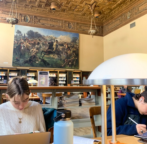 Students studying at a table in the second-floor hall inside of Doe Library. A large painting hangs on the wall in the background behind them. Photo taken by Preslee Vanlandingham.