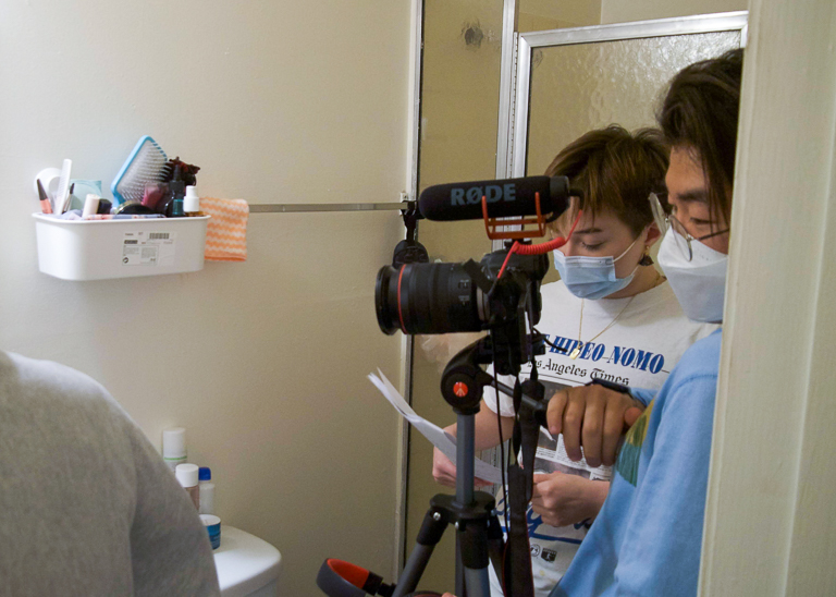 A film set staged in a bathroom. Nina (left) reads a script in the background while Sean (right) operates a DSLR camera mounted on a tripod.