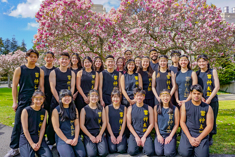 Members of Cal Raijin Taiko pose together in blue and gold uniforms in front of flowering pink trees.