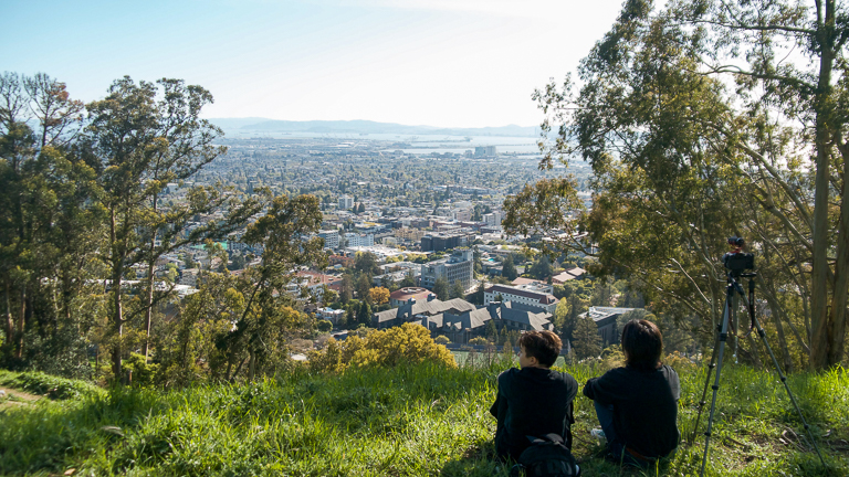 Nina (left) and Sean sit in the Big “C” hills, their backs turned to the camera. The Bay Area skyline can be seen in front of them and tripod to their right.