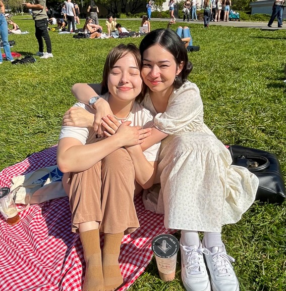 Two students, Nina (left) and Zoe, sit together on a red picnic blanket on Memorial Glade. It’s a sunny, clear day and the Glade is crowded.