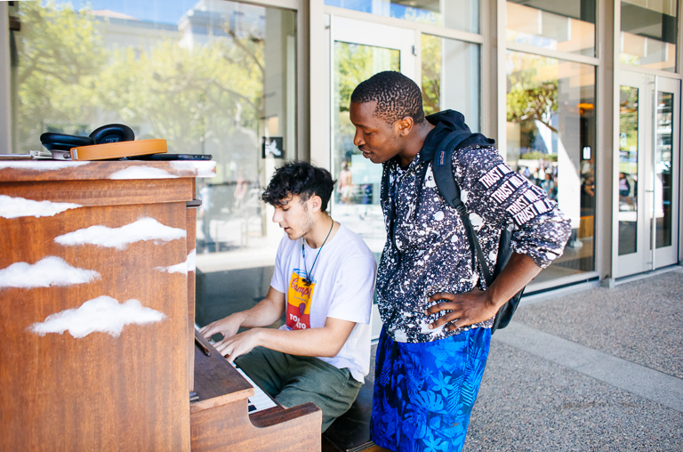 One student sits at the community piano on Sproul in front of the MLK Jr. Building while another student stands next to them watching.