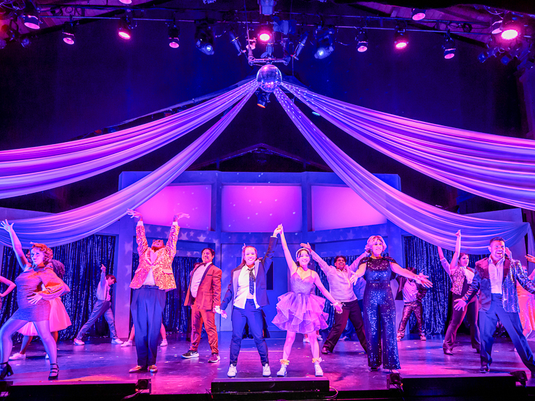 Performers of Prom dance in glittering costumes under purple stage lights.