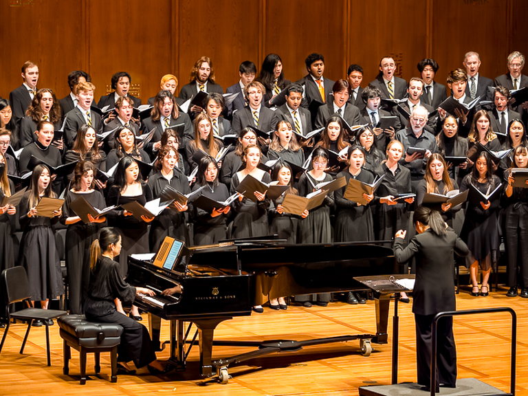 Chamber chorus members perform in all black, guided by a conductor and pianist in front.