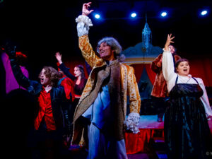 Three actors raise their right hands to the ceiling as they perform on a colorfully illuminated stage in costume.